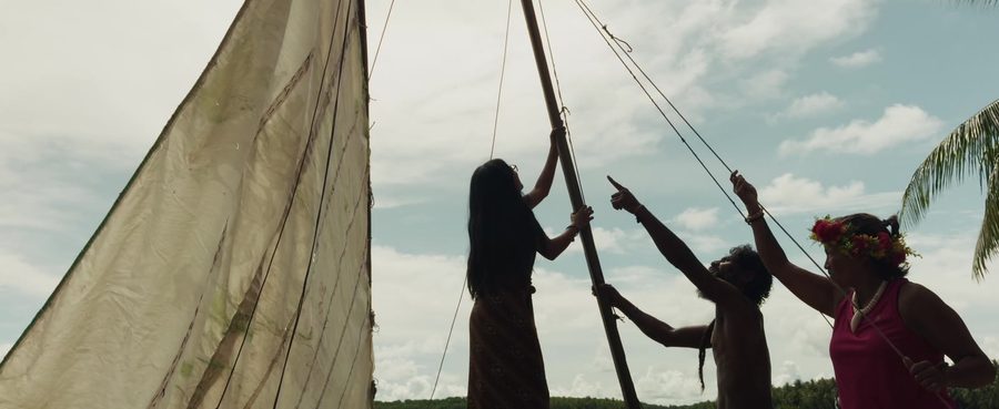 A crew raises a canoe sail against the sky