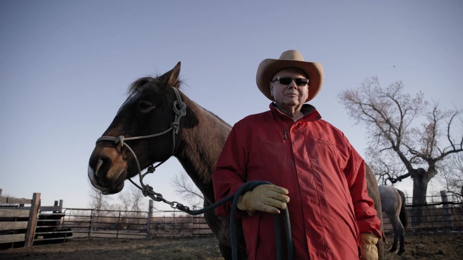 A rancher in a red jacket and cowboy hat stands with his horse
